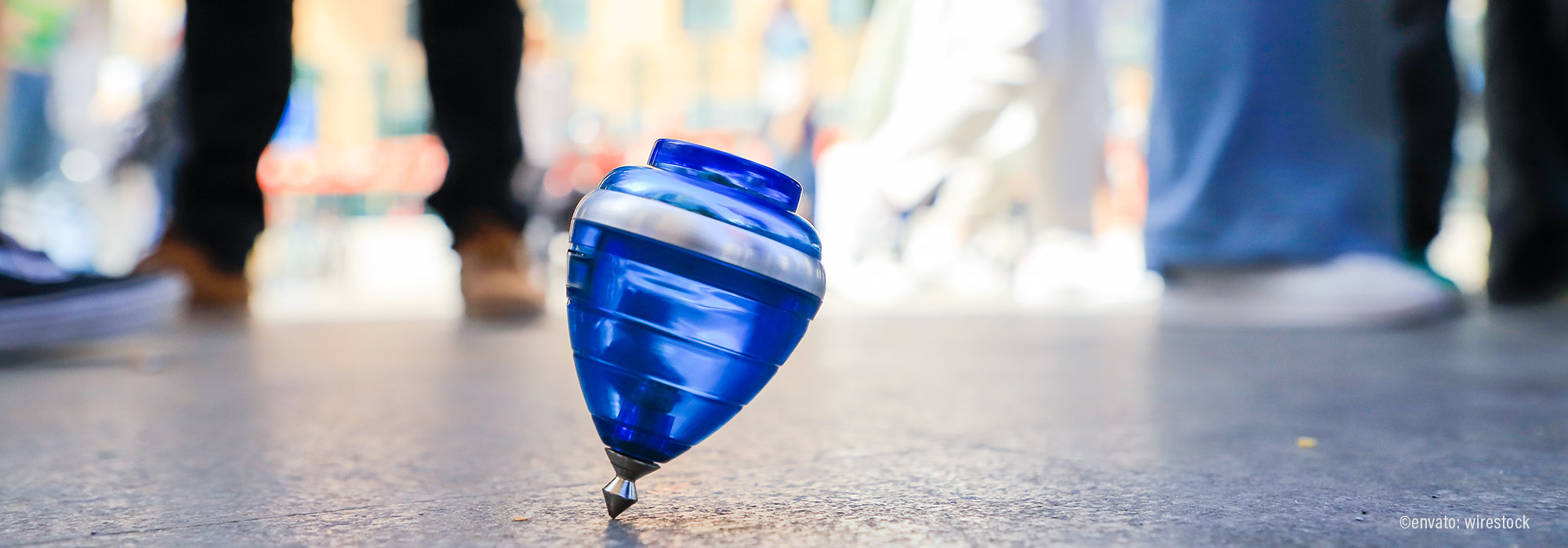 Blau-weißer Kreisel in Drehbewegung auf Boden mit Menschen im Hintergrund - NEBE Impuls Beratung bewegt sich Mut zu kreativen Methoden Lebenshilfe